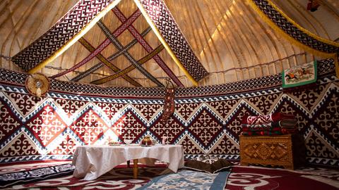 Blick in eine traditionelle Jurte aus Zentralasien, in der ein flacher Tisch mit weißer Tischdecke und eine Holttruhe mit Schnitzereien steht. Auf dem Boden liegen verschiedenfarbige Teppiche.