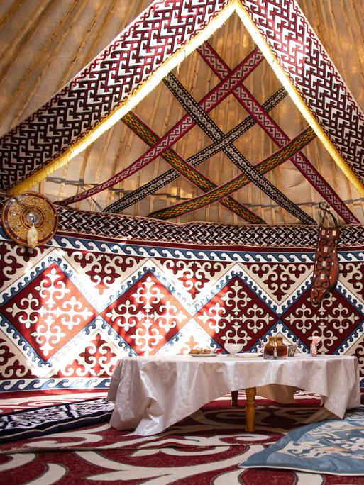 Blick in eine traditionelle Jurte aus Zentralasien, in der ein flacher Tisch mit weißer Tischdecke und eine Holttruhe mit Schnitzereien steht. Auf dem Boden liegen verschiedenfarbige Teppiche.