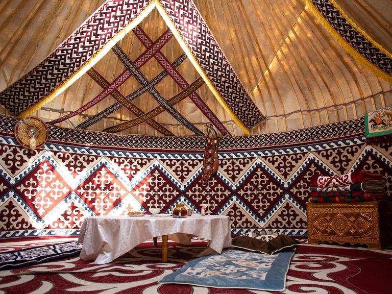 Blick in eine traditionelle Jurte aus Zentralasien, in der ein flacher Tisch mit weißer Tischdecke und eine Holttruhe mit Schnitzereien steht. Auf dem Boden liegen verschiedenfarbige Teppiche.
