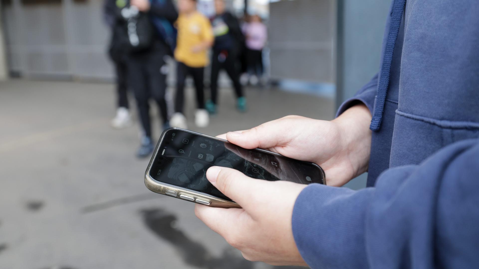 Ein Schüler hält ein Smartphone in der Hand, im Hintergrund sind Jugendliche verschwommen zu sehen