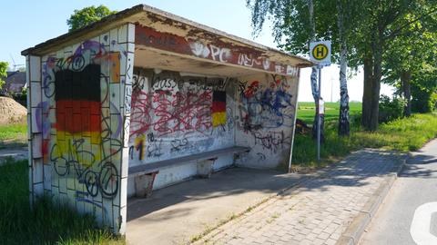 Das Foto zeigt ein steinernes Wartehäuschen einer Bushaltestelle. Das Häuschen ist stark beschmiert und bemalt. Das auffälligste Element: zwei große aufgemalte Deutschlandfahnen. 