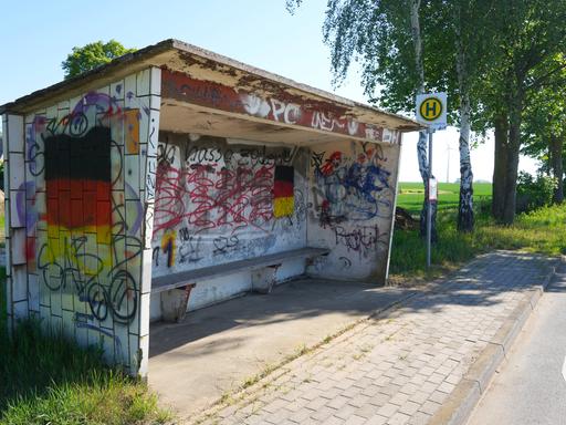 Das Foto zeigt ein steinernes Wartehäuschen einer Bushaltestelle. Das Häuschen ist stark beschmiert und bemalt. Das auffälligste Element: zwei große aufgemalte Deutschlandfahnen. 