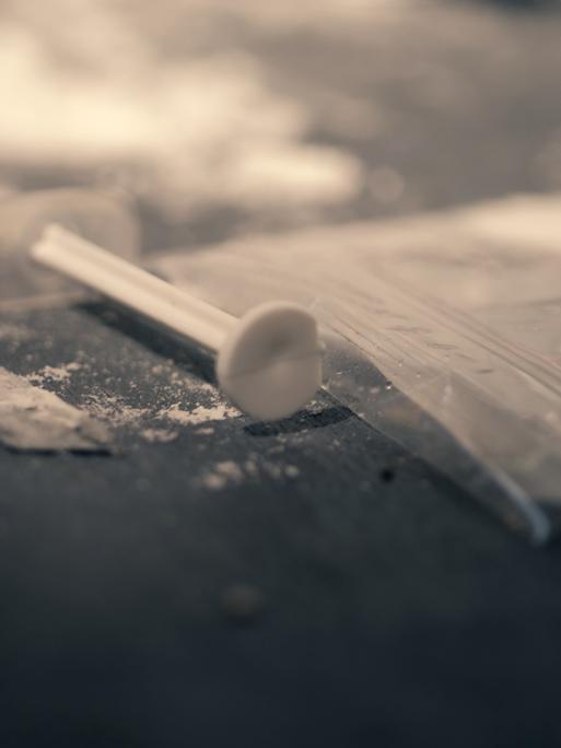 Ein Beutel mit einem weißen Pulver liegt neben einer Spritze. Es soll den Konsum von Heroin andeuten. 