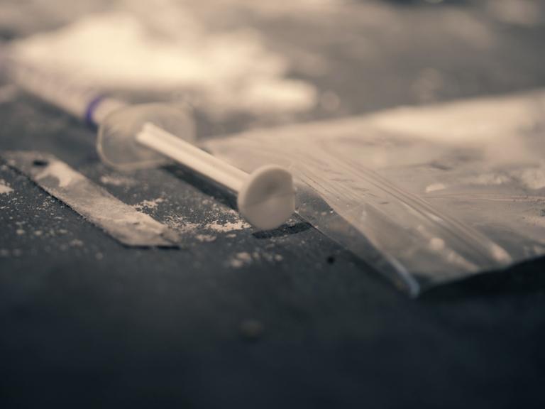 Ein Beutel mit einem weißen Pulver liegt neben einer Spritze. Es soll den Konsum von Heroin andeuten. 