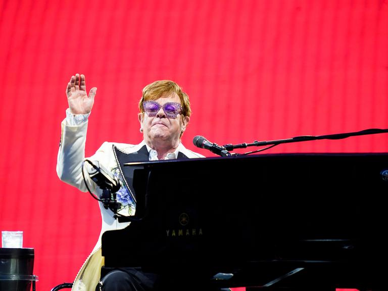 Der Musiker Elton John sitzt am Flügel auf einer Bühne vor einem roten Hintergrund