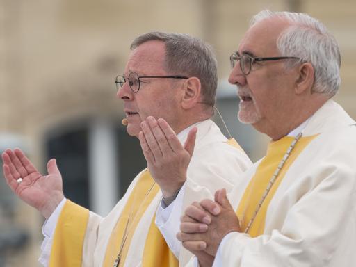 Zwei ältere Brillenträger in weiß-gelben Gewändern nehmen im Freien an einem katholischen Gottesdienst teil.
