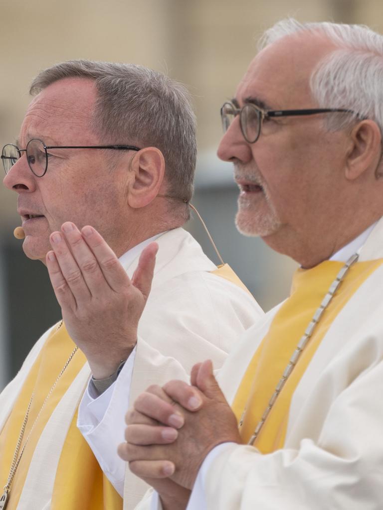 Zwei ältere Brillenträger in weiß-gelben Gewändern nehmen im Freien an einem katholischen Gottesdienst teil.