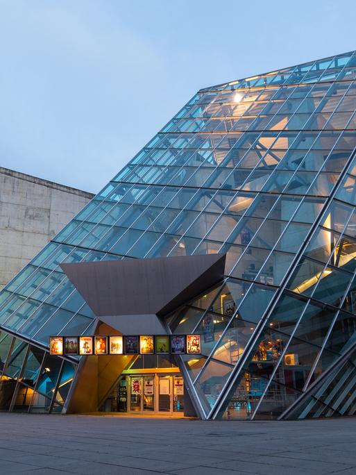 Ein schräges Glasgebäude in der Innnenstadt ist am Abend beleuchtet
