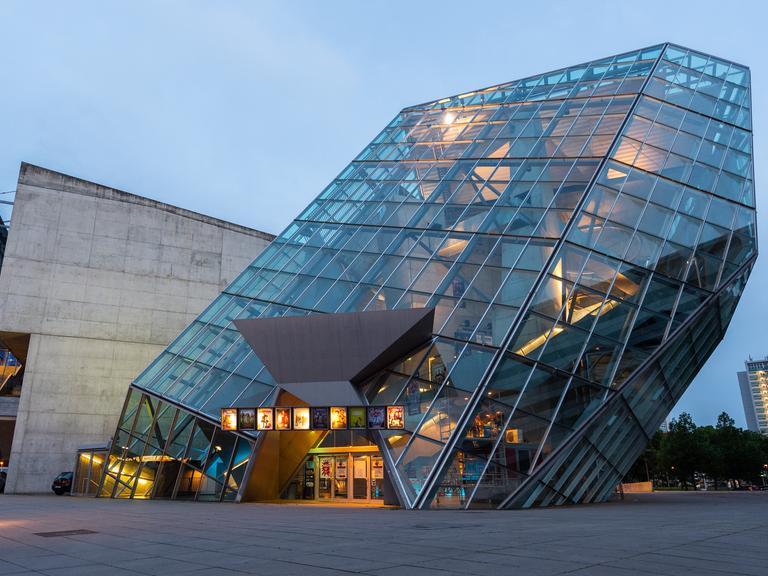 Ein schräges Glasgebäude in der Innnenstadt ist am Abend beleuchtet