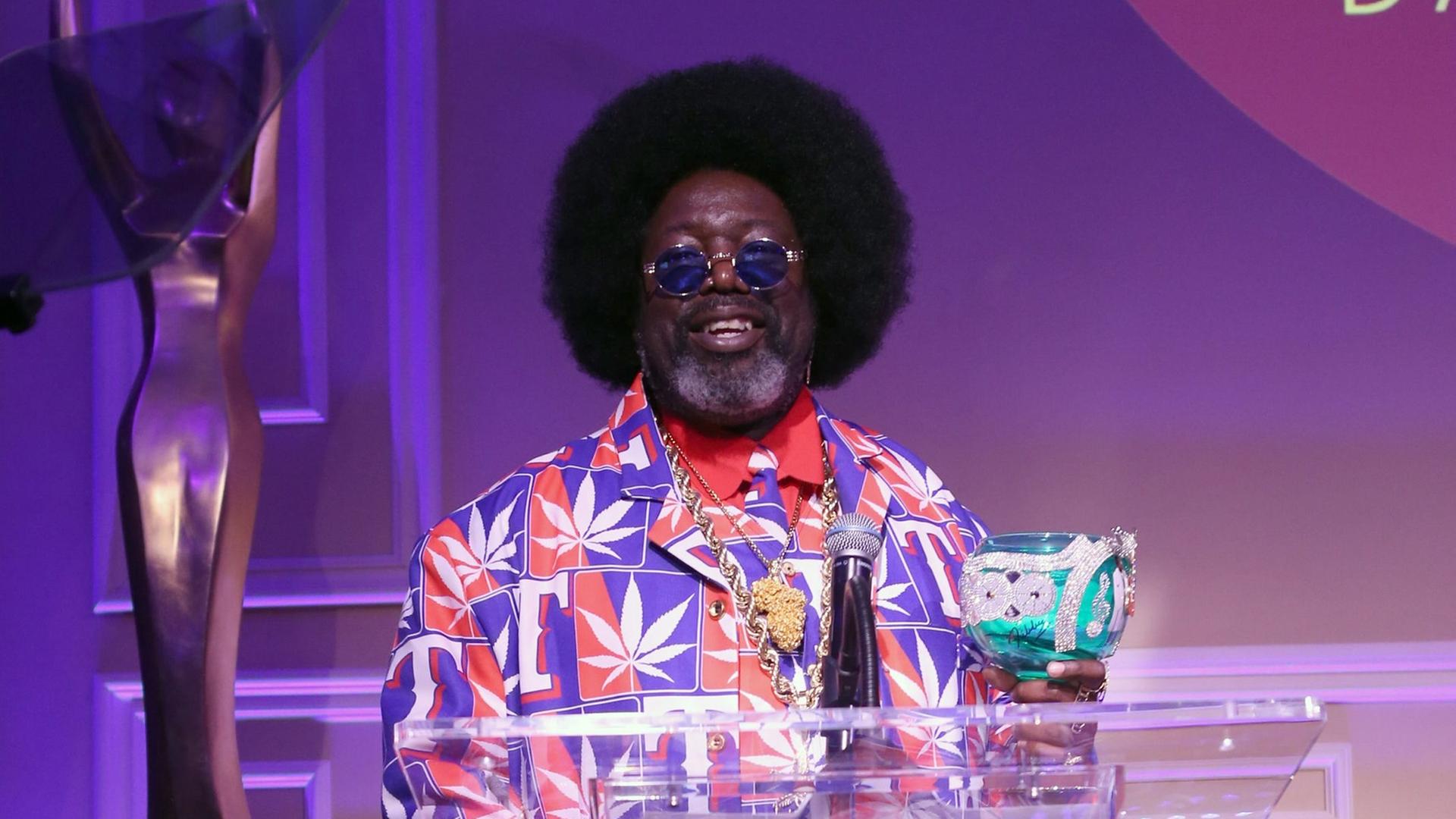 Der Rapper Afroman steht in einem sehr bunten Anzug mit aufgedruckten Cannabis-Blättern hinter einem durchsichtigen Redepult. Er trägt Bart, Sonnenbrille und eine Afro-Frisur.