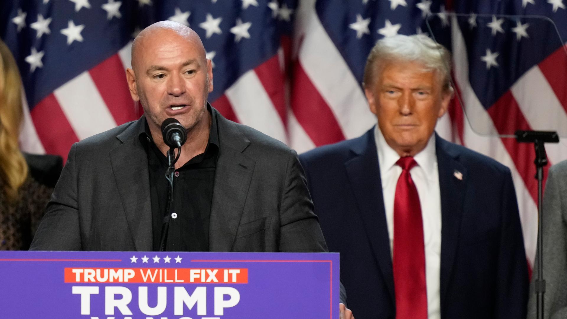 Dana White steht am Rednerpult. Donald Trump steht versetzt dahinter und hört zu.