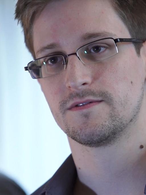 Edward Snowden mit grauem Hemd und Brille sitzt mit leicht schrägem Kopf so, dass sein Kopf sich im hinter ihm stehenden Spiegel spiegelt.