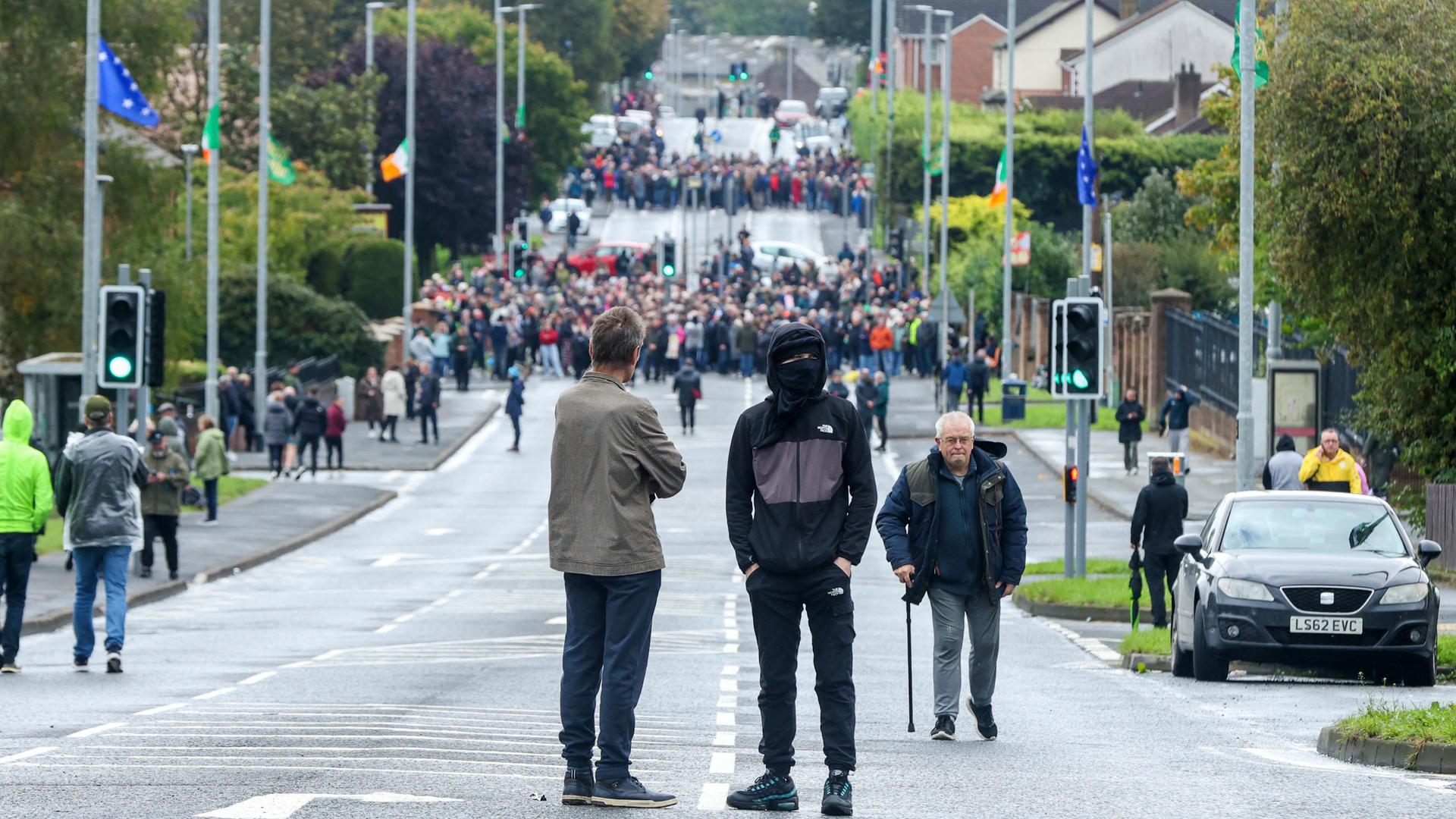 Blick auf eine Straße, auf der mehrere große Menschengruppen die Kreuzungen blockieren. Im Vordergrund steht ein Mann mit Maske und Kaputze. Daneben läuft ein älterer Mann mit Stock entlang.