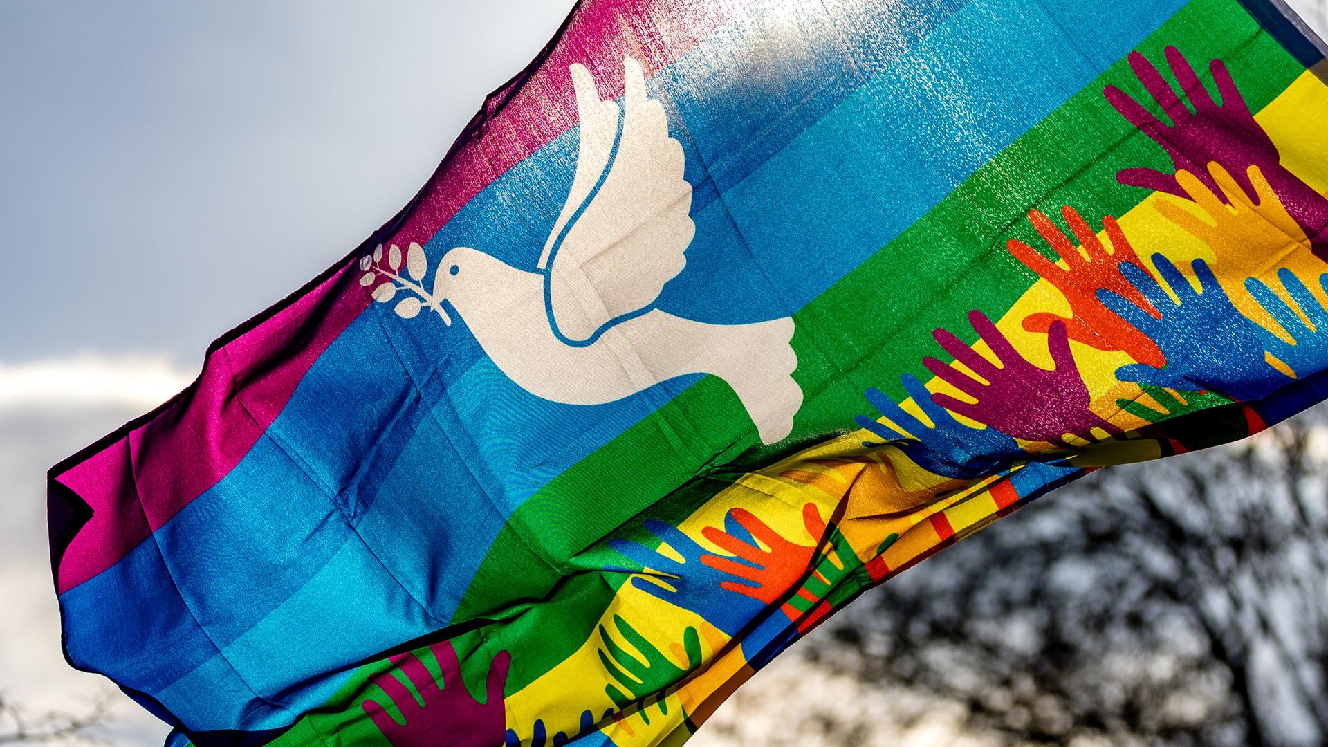 Hände in Regenbogenfarben und eine weiße Friedenstaube sind auf einer Regenbogenfahne bei der Demonstration.