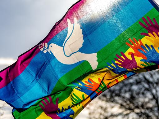 Hände in Regenbogenfarben und eine weiße Friedenstaube sind auf einer Regenbogenfahne bei der Demonstration.