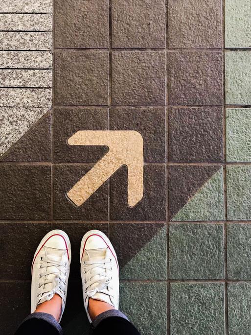 Die Füße einer Frau mit Turnschuhen stehen auf einer Straße, auf der eine Spur und ein Pfeil aufgemalt sind.
