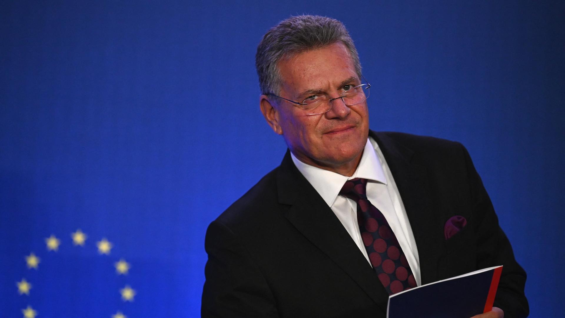 EU-Handelskommissar Sefcovic steht vor einem blauen Hintergrund, darauf ist die EU-Flagge zu sehen. Er trägt einen schwarzen Anzug, ein weißes Hemd und eine rot-gepunktete Krawatte. 
