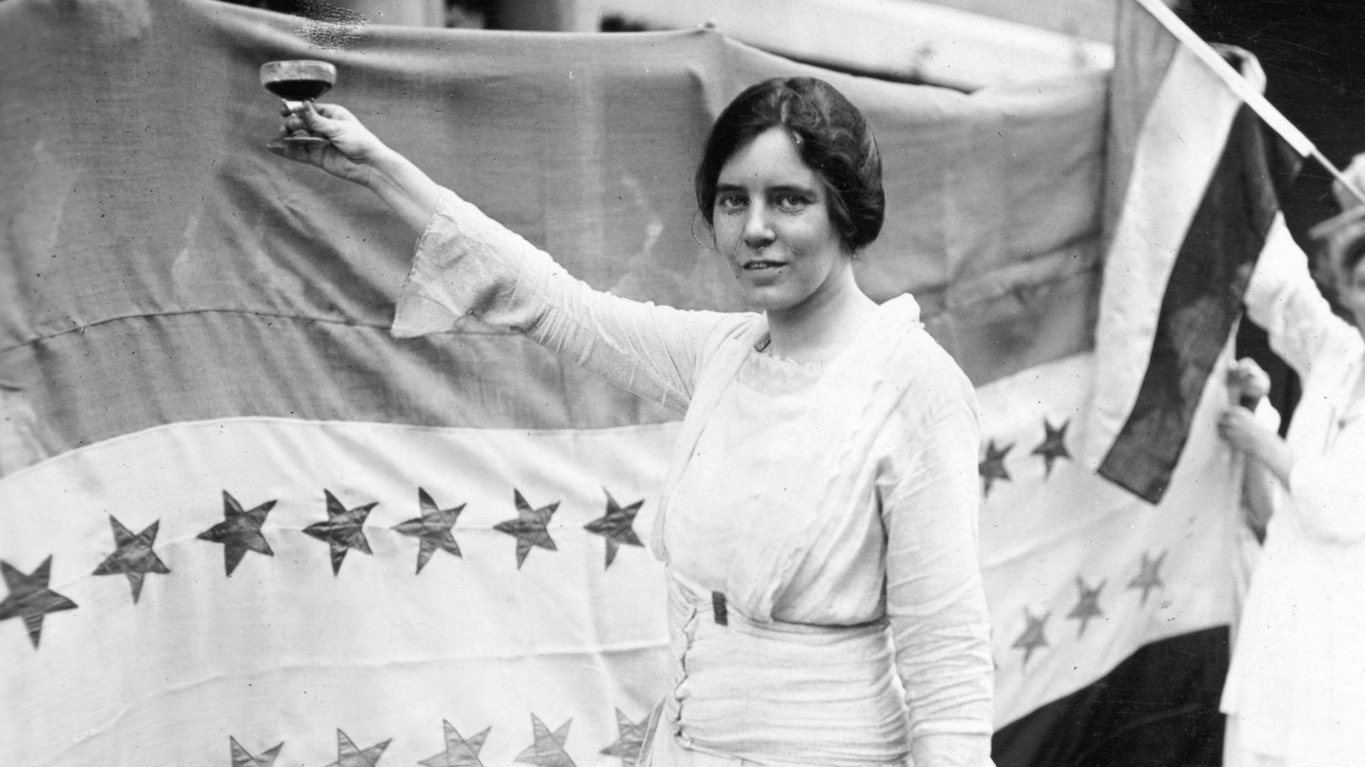 Historisches Schwarzweißfoto von Alice Paul, die ein weißes Kleid trägt und eine Hand mit einem Sektglas erhebt.