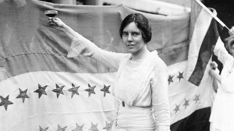 Historisches Schwarzweißfoto von Alice Paul, die ein weißes Kleid trägt und eine Hand mit einem Sektglas erhebt.