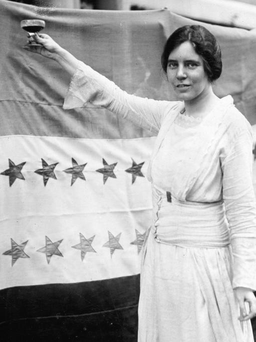 Historisches Schwarzweißfoto von Alice Paul, die ein weißes Kleid trägt und eine Hand mit einem Sektglas erhebt. Historisches Schwarzweißfoto von Alice Paul, die ein weißes Kleid trägt und eine Hand mit einem Sektglas erhebt.