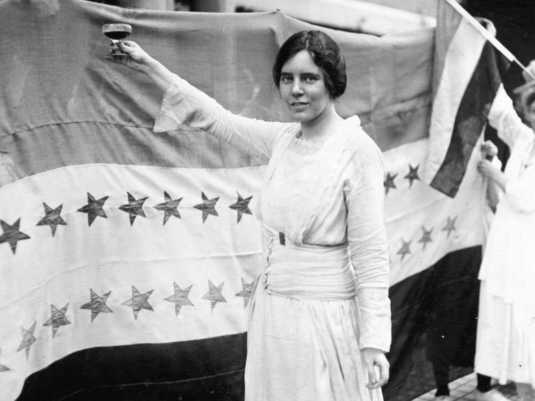 Historisches Schwarzweißfoto von Alice Paul, die ein weißes Kleid trägt und eine Hand mit einem Sektglas erhebt. Historisches Schwarzweißfoto von Alice Paul, die ein weißes Kleid trägt und eine Hand mit einem Sektglas erhebt.