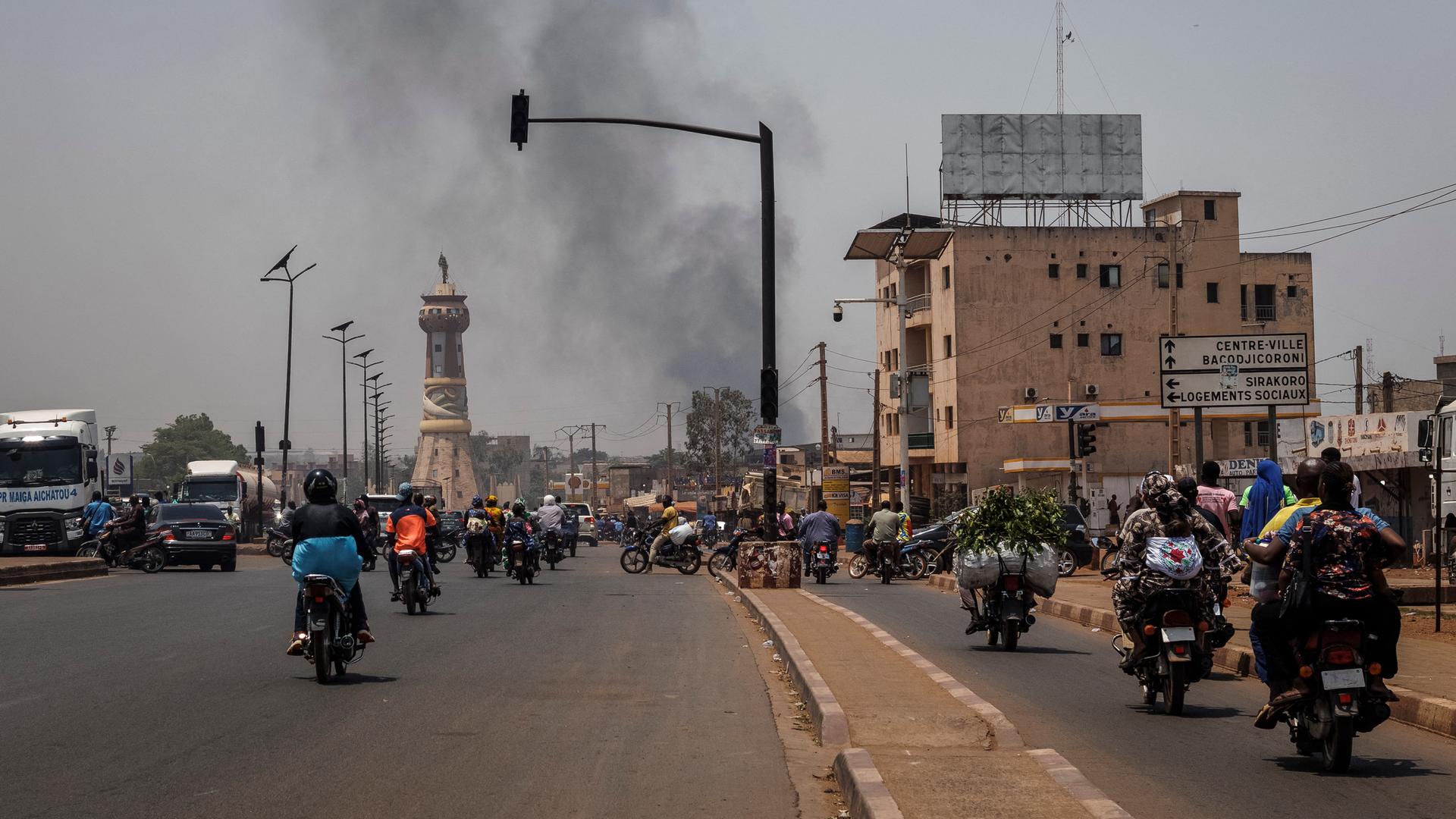 Auf einer Straße fahre viele Motorräder. Im Hintergrund steigt dunkler Rauch auf.