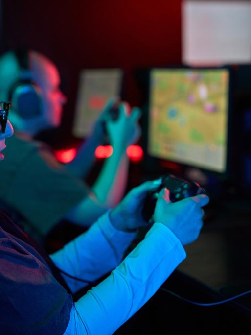 Seitenansicht-Porträt einer Frau, die einen Gaming-Controller hält und Videospiele im Neonlicht spielt. Neben ihr sitzt ein Mann, der auch spielt. Beide tragen Kopfhörer und sind konzentriert.