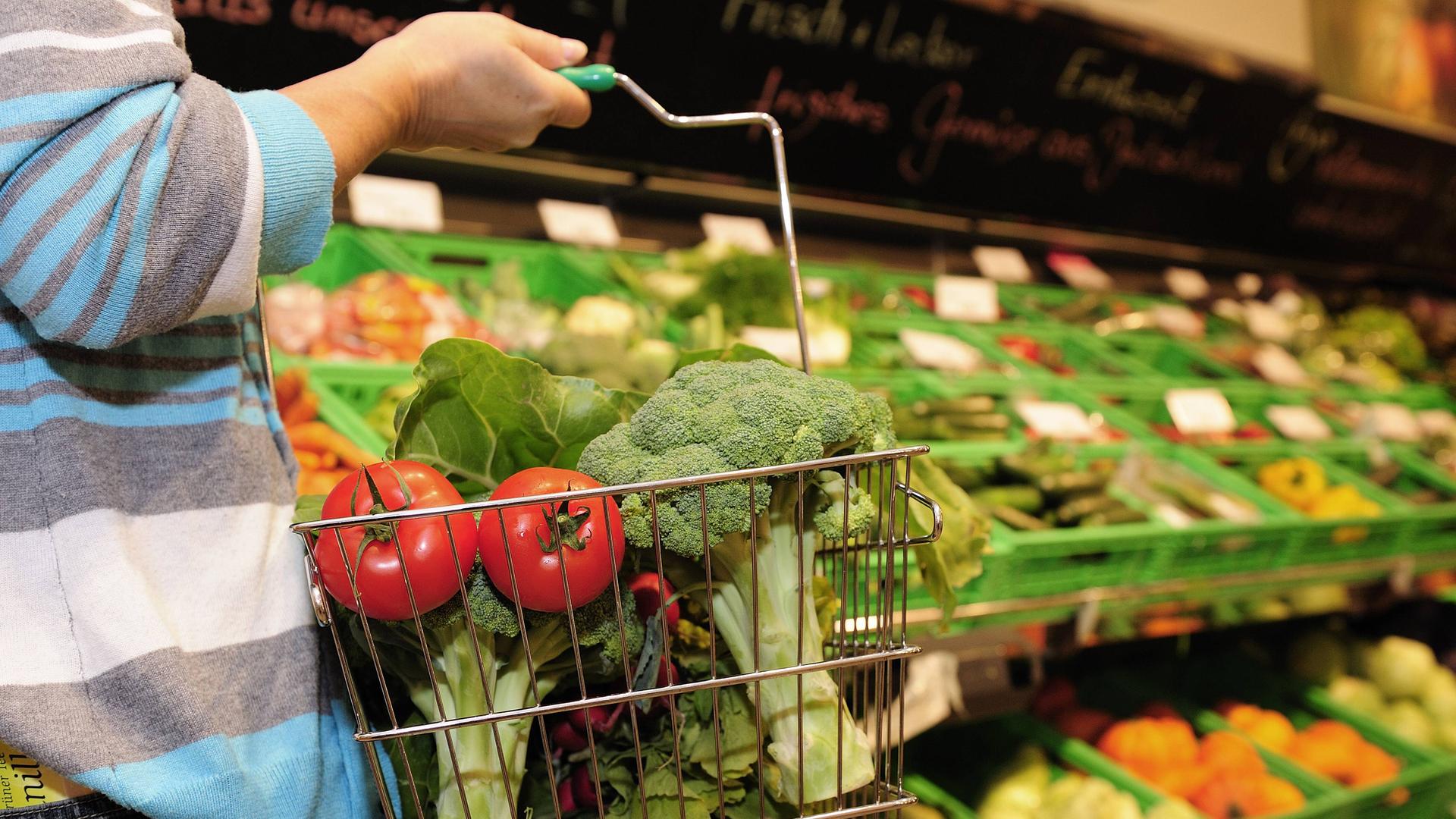Eine Frau läuft durch einen Supermarkt, sie trägt einen Einkaufskorb mit Tomaten und Brokkoli darin. Im Hintergrund steht ein Regal mit Gemüse.
