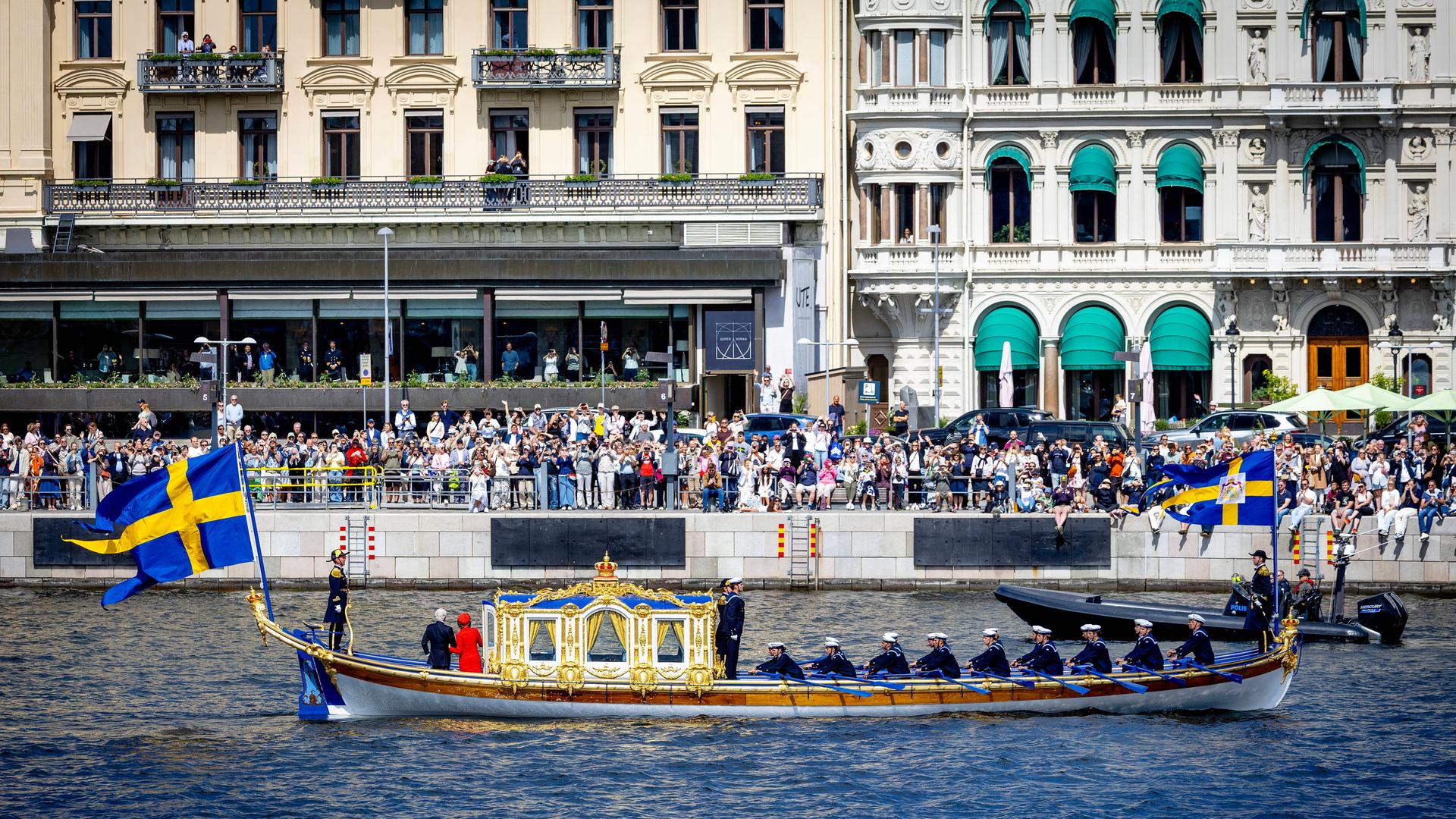Ein Boot fährt über einen Kanal, am Ufer stehen hunderte Menschen und Winken. Am Boot hängt eine Flagge in blau und gelb. Ein Boot fährt über einen Kanal, am Ufer stehen hunderte Menschen und Winken. Am Boot hängt eine Flagge in blau und gelb.
