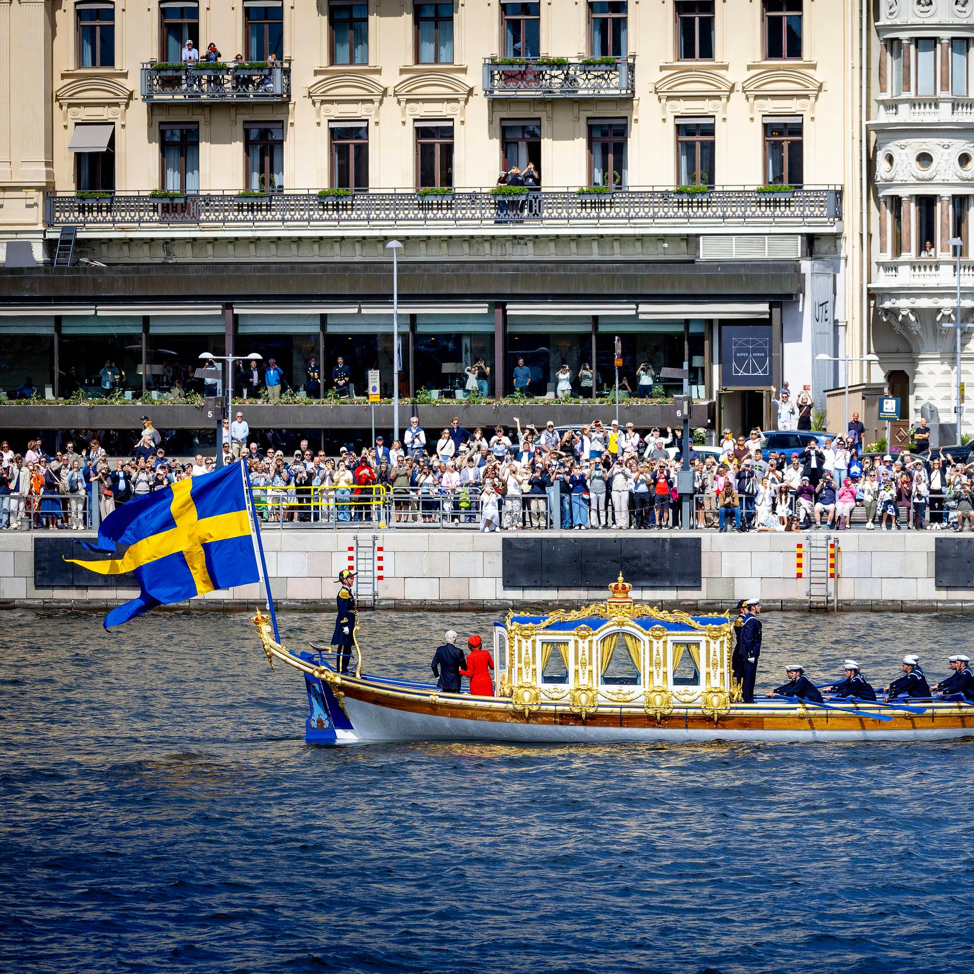 Ein Boot fährt über einen Kanal, am Ufer stehen hunderte Menschen und Winken. Am Boot hängt eine Flagge in blau und gelb.