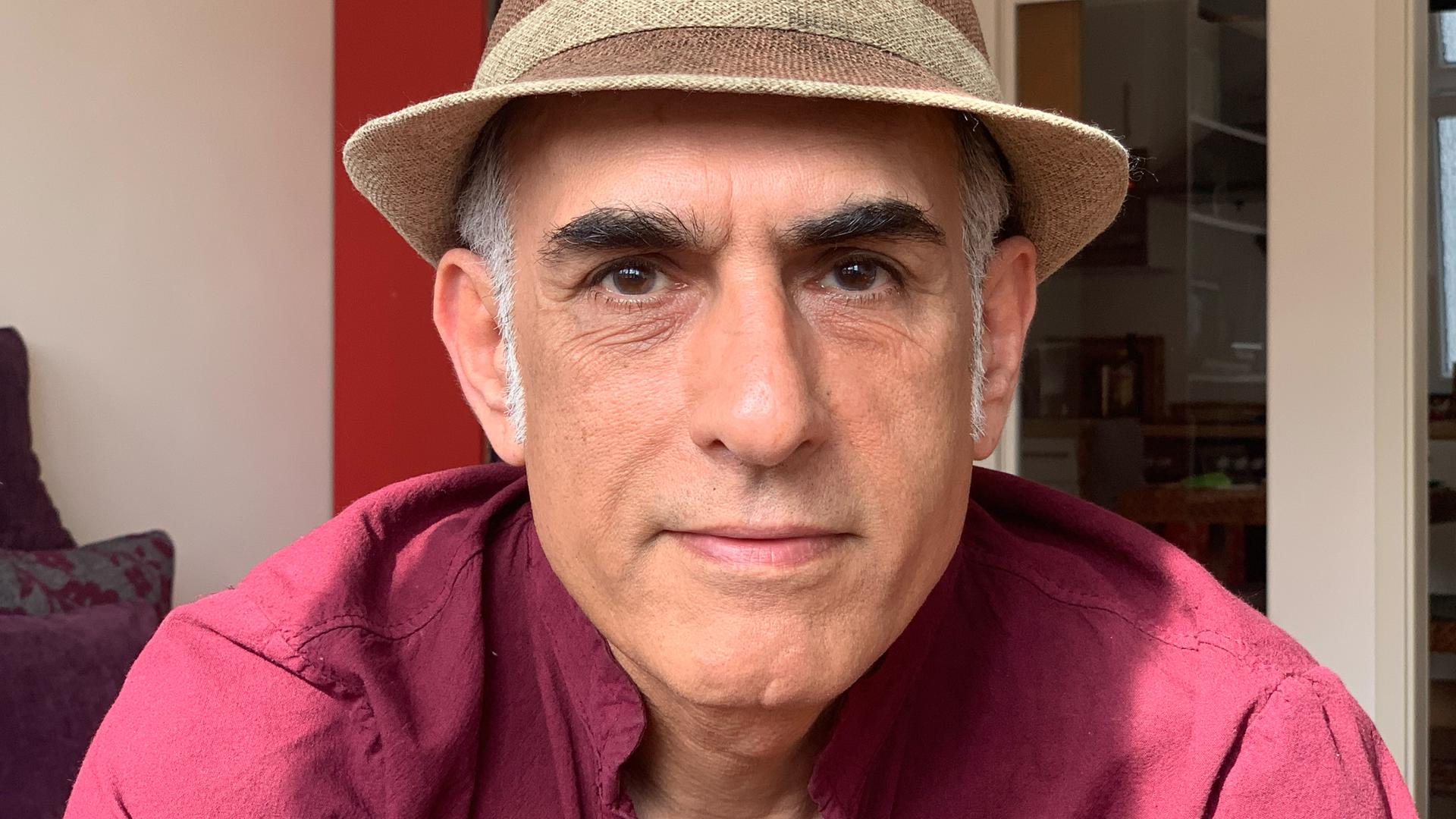 Der iranische Autor Bahram Moradi lächelt in die Kamera. Er trägt ein rotes Hemd und einen braunen Hut.