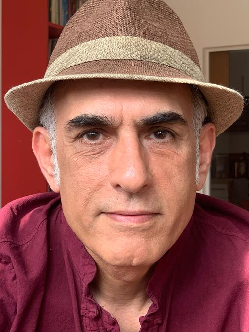 Der iranische Autor Bahram Moradi lächelt in die Kamera. Er trägt ein rotes Hemd und einen braunen Hut. Der iranische Autor Bahram Moradi lächelt in die Kamera. Er trägt ein rotes Hemd und einen braunen Hut.