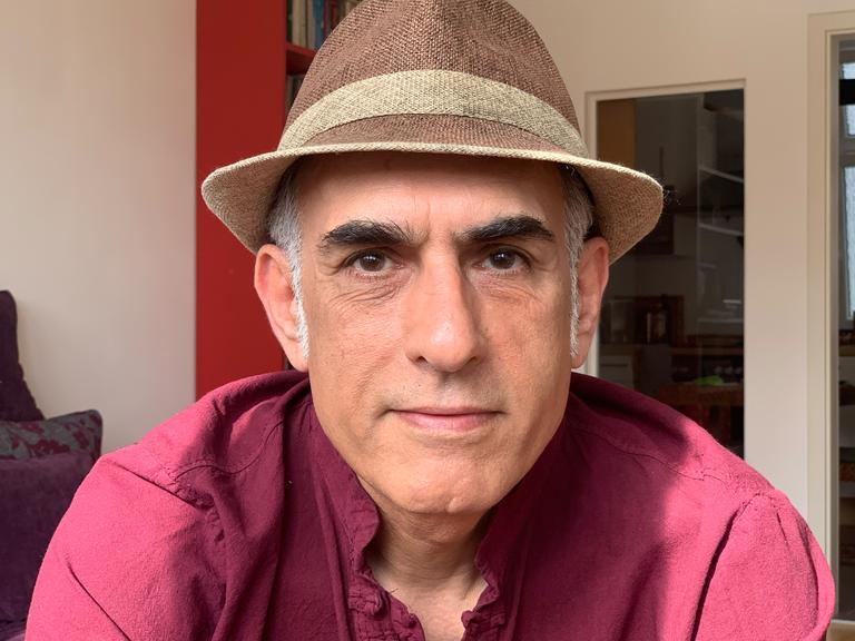 Der iranische Autor Bahram Moradi lächelt in die Kamera. Er trägt ein rotes Hemd und einen braunen Hut. Der iranische Autor Bahram Moradi lächelt in die Kamera. Er trägt ein rotes Hemd und einen braunen Hut.