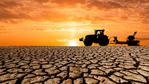 Auf einem völlig ausgetrockneten erdboden ist ein Traktor mit landwirtschaftlichem Anhänger vor untergehender Sonne zu erkennen.