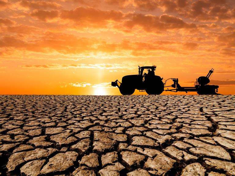 Auf einem völlig ausgetrockneten erdboden ist ein Traktor mit landwirtschaftlichem Anhänger vor untergehender Sonne zu erkennen.