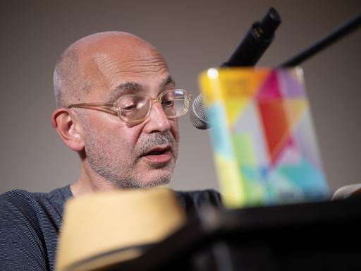 Der Schriftsteller Maxim Billder sitzt auf einer Bühne, trägt Brille und spricht ins Mikrofon