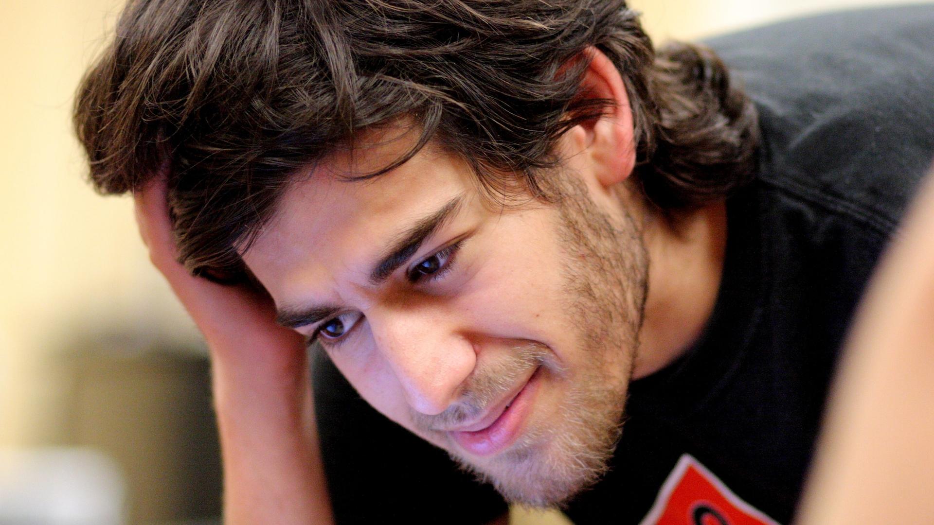 Ein junger Mann mit schwarzen Haaren blickt lächelnd auf einen Bildschirm, er stützt den Kopf auf die rechte Hand.
