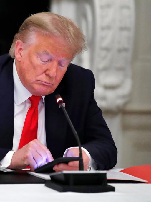 Donald Trump sitz mit gesenktem Blick an einem Konferenztisch und schaut auf das erleuchtete Display seines Smartphones.