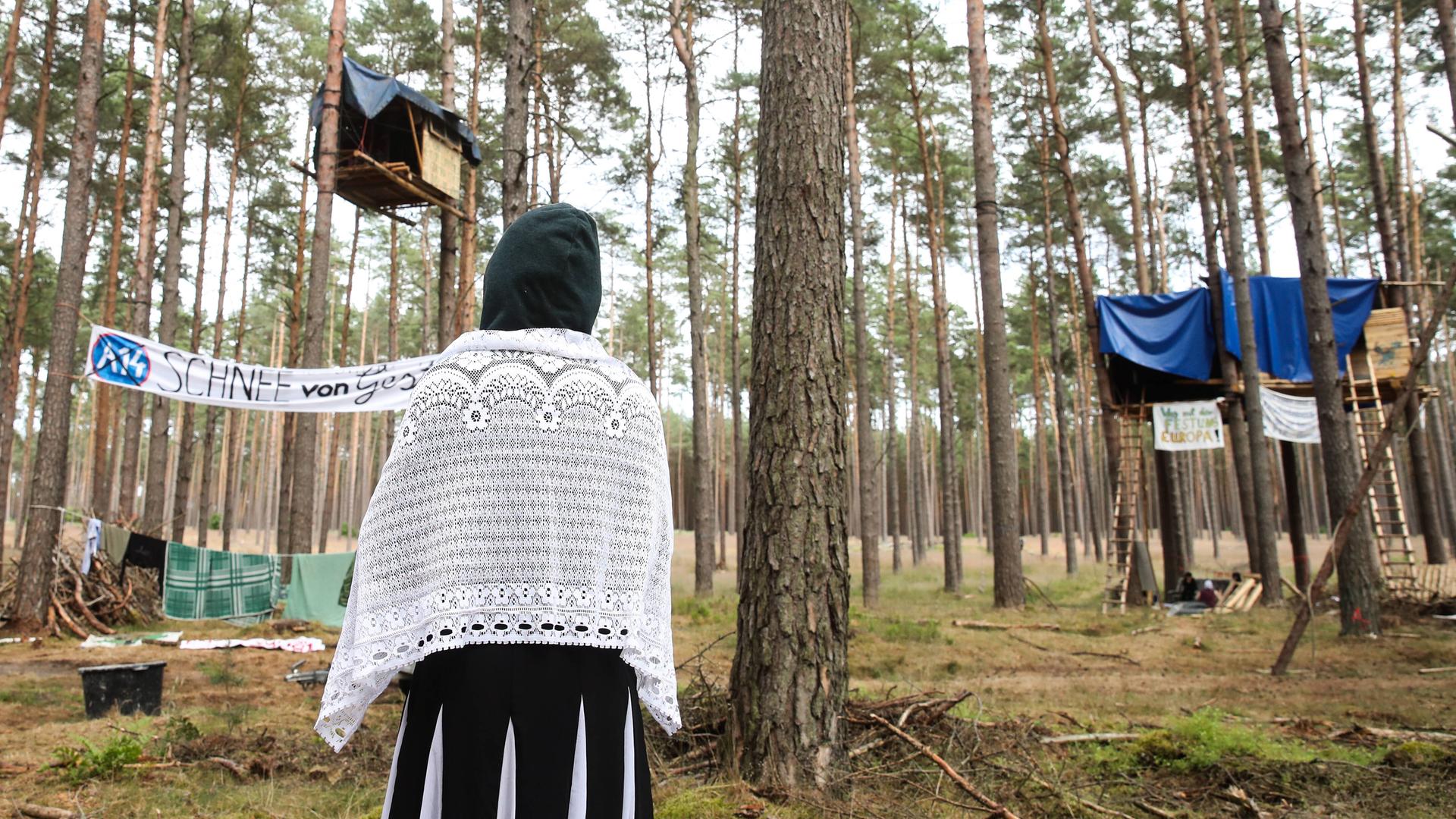 Eine Klimaaktivistin steht mit dem Rücken zur Kamera und schaut auf Baumhäuser als Teil des Protestcamps in einem Kiefernwald bei Seehausen. Auf einem Transparant steht A14 - Schnee von gestern.