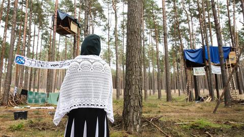 Eine Klimaaktivistin steht mit dem Rücken zur Kamera und schaut auf Baumhäuser als Teil des Protestcamps in einem Kiefernwald bei Seehausen. Auf einem Transparant steht A14 - Schnee von gestern.