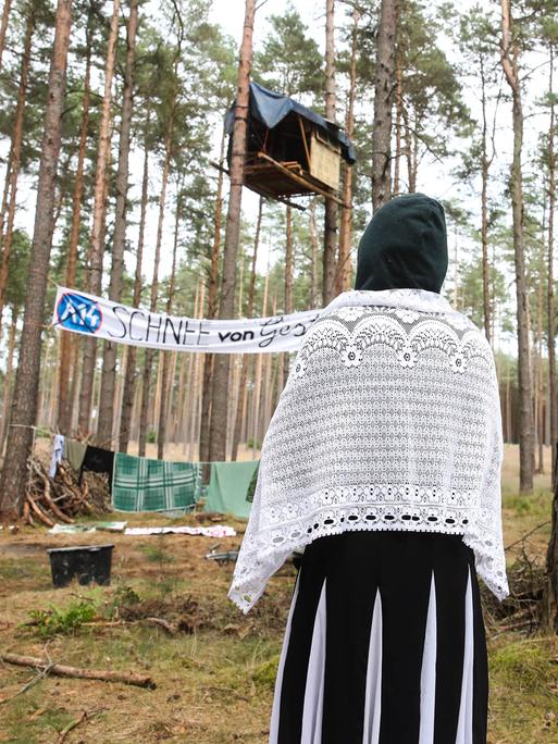 Eine Klimaaktivistin steht mit dem Rücken zur Kamera und schaut auf Baumhäuser als Teil des Protestcamps in einem Kiefernwald bei Seehausen. Auf einem Transparant steht A14 - Schnee von gestern.