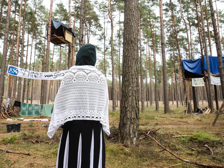 Eine Klimaaktivistin steht mit dem Rücken zur Kamera und schaut auf Baumhäuser als Teil des Protestcamps in einem Kiefernwald bei Seehausen. Auf einem Transparant steht A14 - Schnee von gestern.