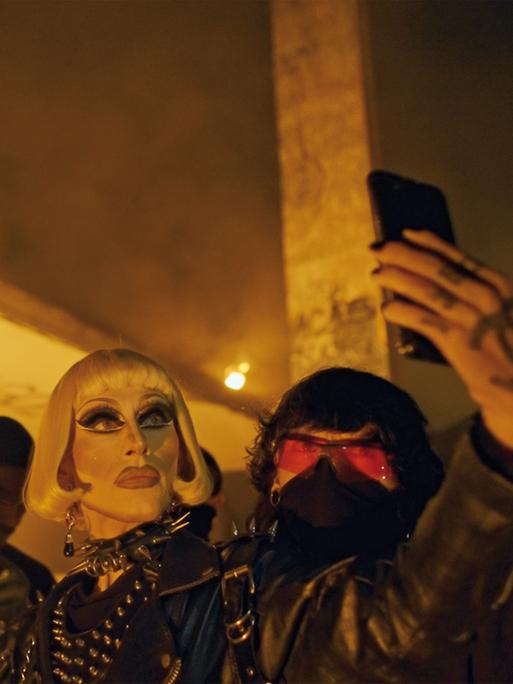 Vermummte und verkleidete queere Menschen stehen nachts vor einem Lagerhaus. Ein geschminkter Mann macht ein Selfie.