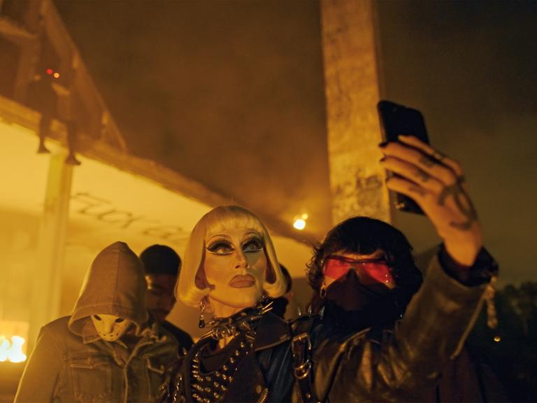 Vermummte und verkleidete queere Menschen stehen nachts vor einem Lagerhaus. Ein geschminkter Mann macht ein Selfie.