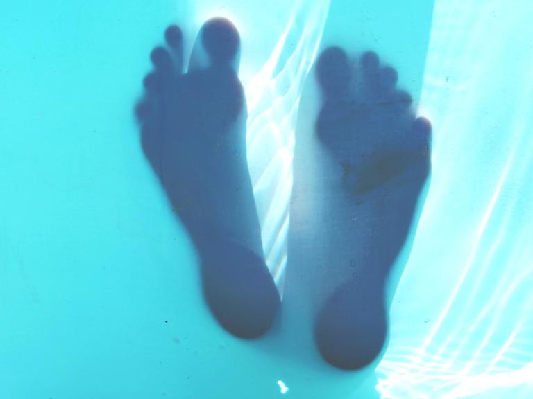 Transparente Unterseite eines Wasserbeckens, auf der sich die Silhouetten zweier Füße abzeichnen.