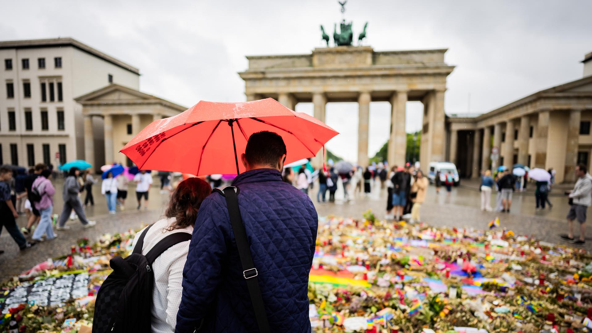 Zwei Personen stehen vor den Trauer- und Anteilbekundungen, die zum Gedenken an die Opfer des Anschlags auf den CSD in Berlin vor dem Brandenburger Tor liegen. Zwei Personen stehen vor den Trauer- und Anteilbekundungen, die zum Gedenken an die Opfer des Anschlags auf den CSD in Berlin vor dem Brandenburger Tor liegen.