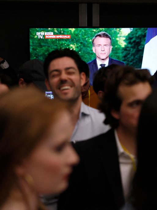 Auf einem TV-Bildschirm verkündet Emmanuel Macron Neuwahlen für Frankreich. Im Vordergrund stehen Mitglieder der rechten Reconquête-Partei und reagieren, teils mit ernsten Gesichtern, teils lachend.