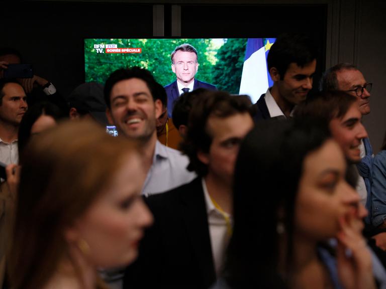 Auf einem TV-Bildschirm verkündet Emmanuel Macron Neuwahlen für Frankreich. Im Vordergrund stehen Mitglieder der rechten Reconquête-Partei und reagieren, teils mit ernsten Gesichtern, teils lachend.