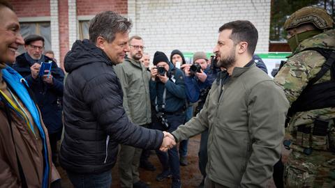 Die beiden schütteln sich lächelnd die Hand; sie sind von der Seite vor einer Hauswand fotografiert. Um sie herum Fotografen und andere Personen sowie ein Soldat in Kampfmonitur.