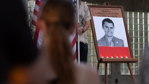Ein Schwarz-Weiß-Foto on Charlie Kirk bei einer Trauerfeier, daneben die US-Flagge, im Vordergrund Menschen
